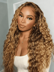 A woman with long, voluminous, wavy blonde highlighted wig wears gold hoop earrings and a gold necklace. She is posing indoors against a light-colored wall.