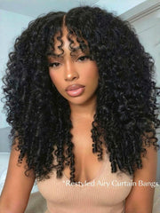 A woman with voluminous, defined curly hair and restyled wispy bangs poses indoors, showcasing a gorgeous Burmese curly wig. She wears a beige top and natural makeup.